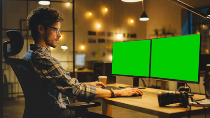 Late Evening in Creative Office: Professional Photographer Works on a Desktop Computer with Two Green Mock-up Screens. Modern Studio Office with Hanging Lightbulbs