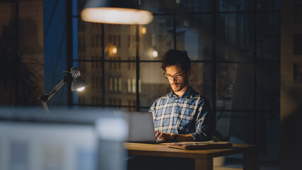 Poster - Handsome Male Creative Office Employee Sitting at His Desk Works on the Laptop. Evening in the Stylish Office Studio with Big Cityscape Window 