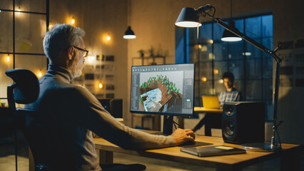 Wall Mural - In the Evening Creative Middle Aged Video Game Developer Works on a Desktop Computer with Screen 3D Videogame Level Design. Professional Office Employee Working Late in the Evening in His Studio 