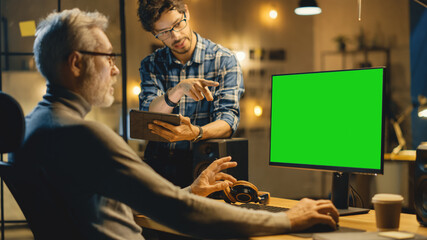 Poster - In the Evening Creative Middle Aged Man Works on a Desktop Computer Green Mock-up Screen, Has Conversation with Younger Specialist who Uses Tablet Computer. Stylish Big City Office late in the Evening
