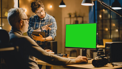 Poster - In the Evening Creative Middle Aged Man Works on a Desktop Computer Green Mock-up Screen, Has Conversation with Younger Specialist who Uses Tablet Computer. Stylish Big City Office late in the Evening