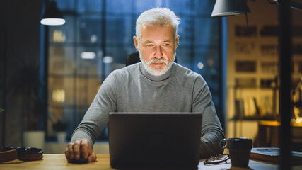 Poster - Portrait of the Handsome and Successful Middle Aged Bearded Businessman Working at His Desk Using Laptop Computer. Working from Cozy Home Office / Studio with Window View of the City at Night