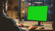 © Gorodenkoff - Over the Shoulder: Creative Young Man Sitting at His Desk Using Desktop Computer with Mock-up Green Screen. Evening in the Stylish Office Studio with City Window View