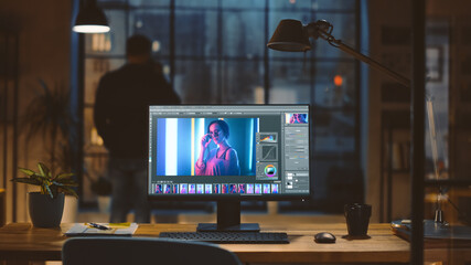 Poster - Desktop Computer with Photo Editing Software Standing on the Desk in the Modern Creative Office. In the Background Designer Drinks from a Cup Looks at the Night City out of the Window.