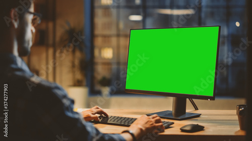 Fotomural Over the Shoulder: Creative Young Man Sitting at His Desk Using Desktop Computer with Mock-up Green Screen