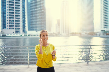 Wall Mural - Enjoying the sound. Happy young woman with earphones listening music on smartphone while walking on city embankment.