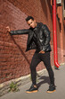 Young handsome stylish guy dancer in a black leather jacket on the street near a red brick wall.