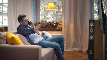 Side view of young man talking on smartphone and watching tv at modern apartment