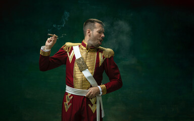 Wall Mural - Disguasted smoking cigarette. Young man in suit as Nicholas II on dark green background. Retro style, comparison of eras concept. Beautiful male model like historical character, monarch, old-fashioned