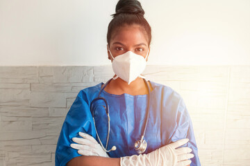 A young African nurse wears a blue uniform, a mask and gloves while standing with her arms crossed in the hospital.