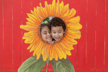 Sister And Brother Try To Squeeze Into The Opening Of A Sunflower Cutout Painting For A Photo Op. The Colorful Sunflower Painted On Plywood With A Red Backdrop.
