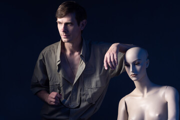 Poster - Portrait of man next to a mannequin. Man with glasses in his hands on a dark background. Man next to a humanoid cyborg. Human-like cyborg.as a symbol of artificial intelligence. Human leans on cyborg