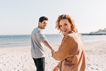 Jocund white girl walking around beach with boyfriend. Handsome brunette man holding hands with girlfriend while posing at sea.
