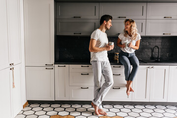 Good-humoured young man drinking tea in kitchen with stylish interior. Indoor portrait of carefree couple enjoying breakfast.