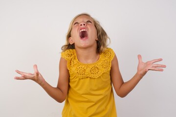 Young blonde kid girl wearing yellow dress over white background crazy and mad shouting and yelling with aggressive expression and arms raised. Frustration concept.