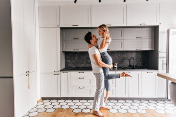 Inspired brunette man holding his wife during indoor photoshoot. Positive couple having fun in kitchen.
