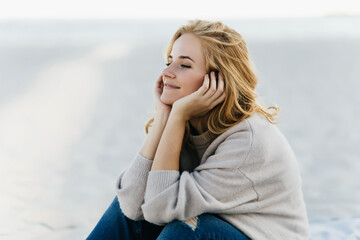 Pensive fair-haired woman chilling at sea background. Adorable cute girl sitting on sandy beach