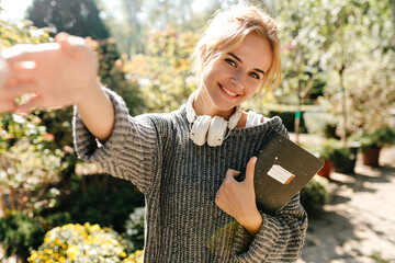 Wall Mural - Photo of green-eyed girl student in gray sweater and headphones, stretching hand for greeting. Lady posing with notepad in park