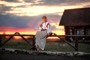 Wall Mural - A beautiful girl in a traditional Russian dress is sitting on a fence against the sunset. The young girl thought about something. Traditional Russian costume.