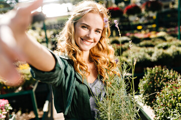 Wall Mural - Portrait of radiant young woman in dark green outfit reaching for camera. Green-eyed model with curls of red color holds lavender bush