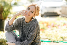 Cute Girl In Knitted Sweater And Gray Jeans Sitting On Bench In Garden And Smiling