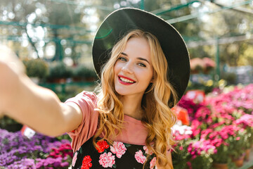 Wall Mural - Adorable white girl taking picture of herself in glasshouse with flowers. Laughing pleasant woman making selfie in orangery