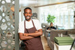 © pressmaster - Cheerful young African waiter in workwear leaning against door in restaurant