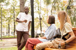 © pressmaster - Happy young African man with drink standing in front of girls relaxing on bench