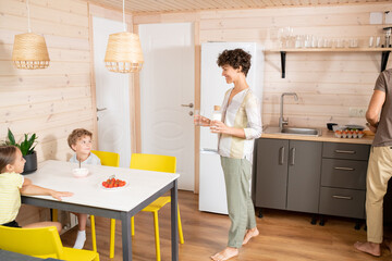 Sticker - Happy young woman with bottle of milk and glass moving towards her kids