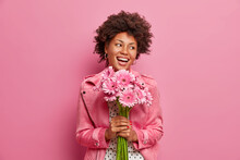 Happy Sincere Afro American Birthday Girl Receives Flowers From Friends, Holds Bouquet Of Gerberas, Has Positive Festive Mood, Isolated On Pink Wall. Glad Woman Accepts Proposal Of Marriage.