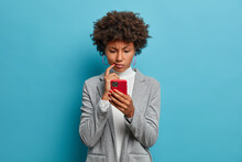 Puzzled Confused Dark Skinned Woman In Formal Wear Looks Indignant At Smartphone, Gazes At Display, Reads Business News On Website, Isolated Blue Background. People And Modern Technology Concept