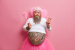© wayhome.studio  - Plump bearded childish male fairy has fun on costume party, dressed in pink pleated skirt, wears wings, has ability to fly, holds magic wand, makes victory gesture, poses against rosy background.
