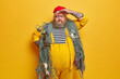 © wayhome.studio  - Bearded serious fisher keeps hand on forehead and looks into distance, poses with fishing net over shoulders, catches sea creatures, has big belly, tattooed arms, stands against yellow background