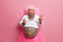 Plump Bearded Childish Male Fairy Has Fun On Costume Party, Dressed In Pink Pleated Skirt, Wears Wings, Has Ability To Fly, Holds Magic Wand, Makes Victory Gesture, Poses Against Rosy Background.