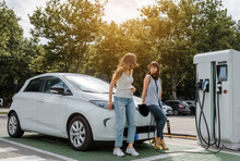 Two Happy Young Beautiful Women Are Talking To Each Other Until Their Electric Car Is Charging At The Charging Station Situated In The Car Park.