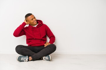 Young latin man sitting on the floor isolated touching back of head, thinking and making a choice.