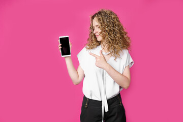 Look at this cell phone. Contented happy woman, pointing her index finger at a blank screen, shows a modern device. Isolated on a pink background.