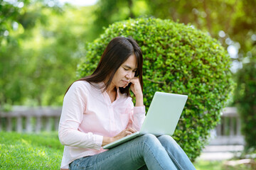 Asian woman sitting green park using laptop computer. Woman working on laptop happy entrepreneur business using notebook with hands typing on keyboard home office during coronavirus quarantine period