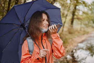 Young caucasian woman in a coronavirus mask holding an umbrella using a smartphone in the autumn forest on a cloudy day. A woman records a voice message on a smartphone in the autumn forest.