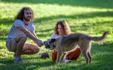 Wall Mural - Young beautiful curly couple man and woman playing with their dog in the park on green grass