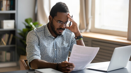 Stressed dissatisfied African American businessman reading letter with bad news, unexpected debt, bank or job dismiss notification, student working on difficult project, holding document in hand