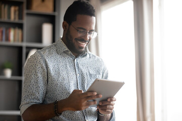 smiling african american man wearing glasses holding computer tablet, looking at screen, playing gam