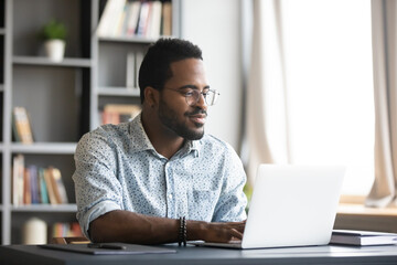 satisfied african american man wearing glasses looking at computer screen, reading good news in emai