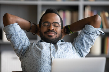 Close up calm African American businessman relaxing daydreaming at workplace, satisfied young man sitting with hands behind head, leaning back in comfortable office chair with closed eyes