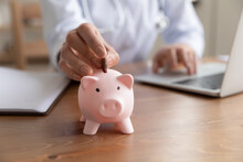 Close Up Young Woman Doctor Putting Coin Into Pink Piggy Bank, Sitting At Work Desk, Using Laptop, Hospital Budget And Accounting, Finances, Medical Insurance, Healthcare Money Savings Concept
