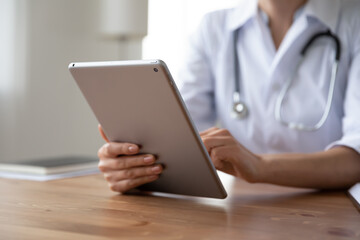 Wall Mural - Close up young woman doctor holding digital tablet, working online, sitting at desk, female physician wearing white coat with stethoscope using medical apps, consulting patient, telemedicine