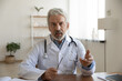 © fizkes - Head shot serious mature doctor wearing white uniform coat with stethoscope speaking, looking at camera, gesturing, senior therapist practitioner gp consulting patient online, making video call