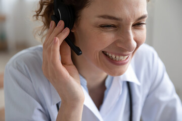 Wall Mural - Close up smiling young woman doctor wearing headset with microphone working online, friendly therapist physician gp wearing white coat uniform consulting patient, telemedicine service, video call