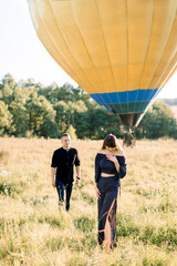 Happy charming young woman in fashionable black clothes, standing in beautiful summer green field, while her boyfriend is walking to her. Hot yellow air balloon on the background