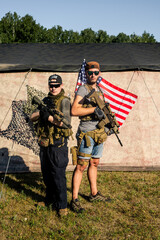 Wall Mural - Portrait of serious young friends in caps and sunglasses holding rifles against military tent with American flag